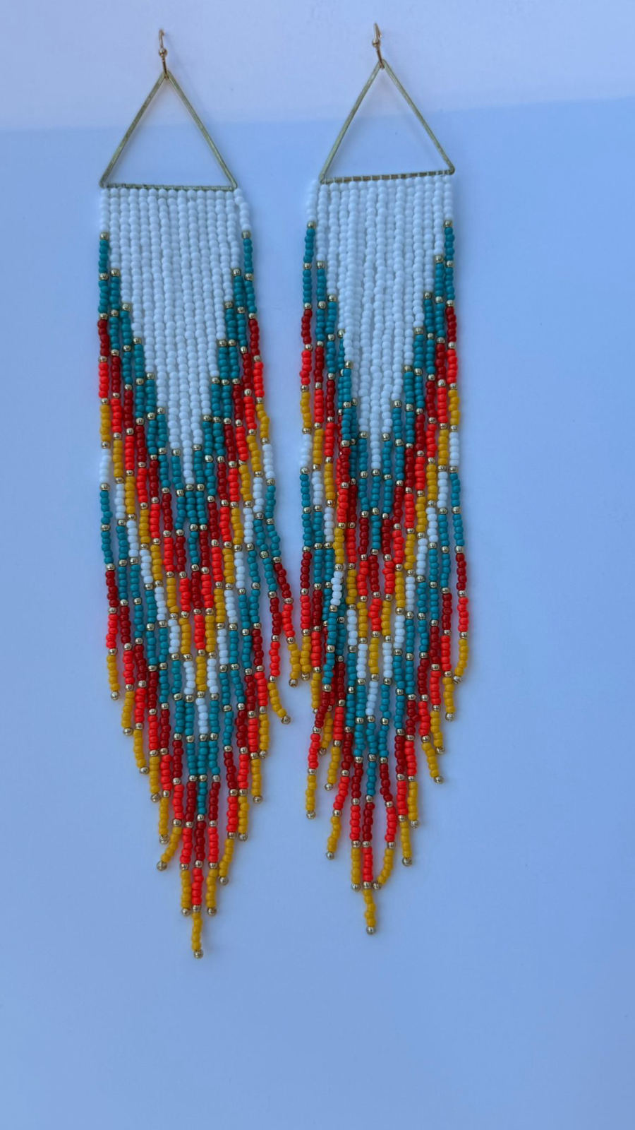 Long fringe drop earrings suspended from open gold triangular metal frames, dense white seed bead fringe patterned with teal, tomato red, and golden yellow in a diagonal flame wave motif, gold metallic round accent beads throughout, pale blue-grey background.